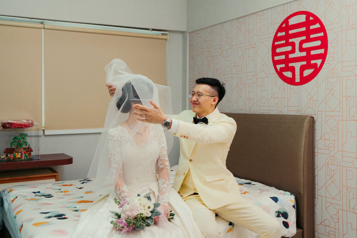 Groom in cream suit lifting bride's veil in decorated wedding room with red double happiness symbol.