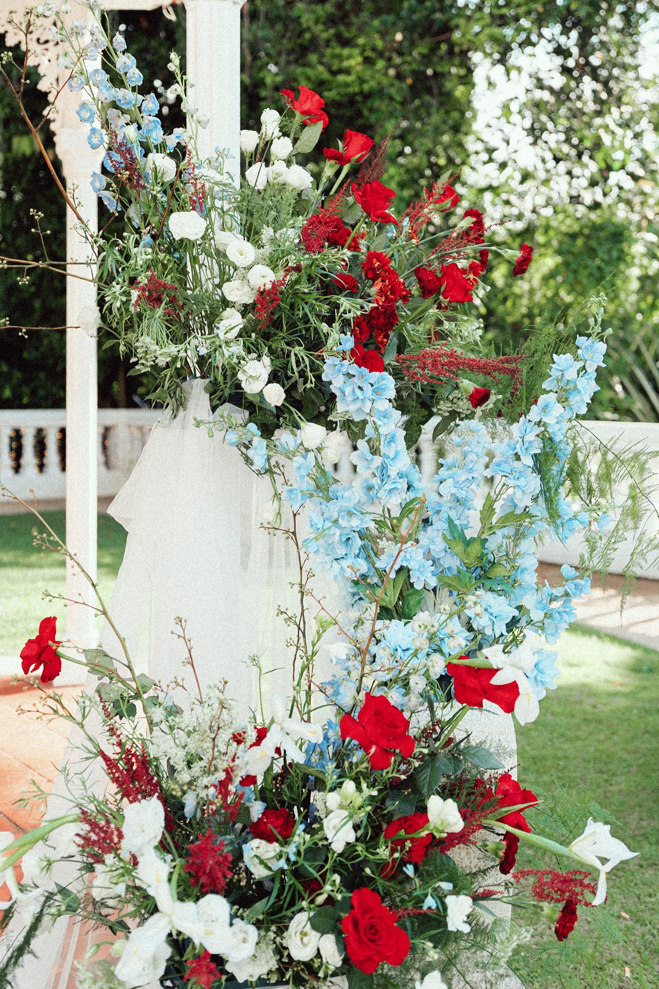 Elegant floral arrangement featuring red carnations, white roses and light blue delphinium flowers against a garden backdrop.