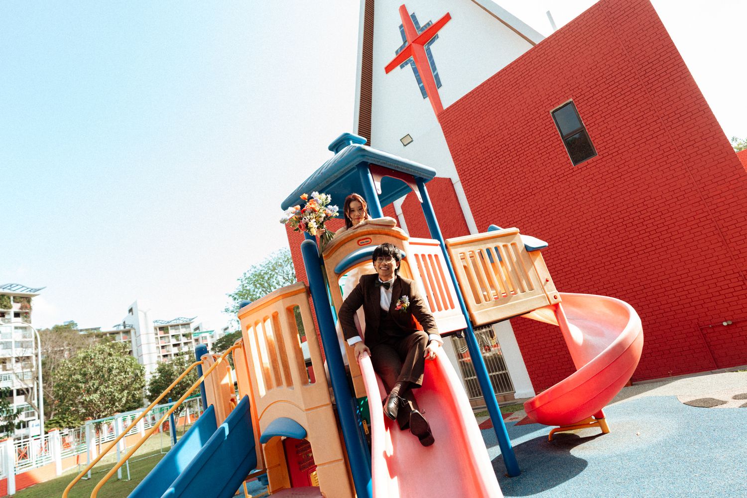 A man in formal attire sits on a colorful playground slide at a vibrant outdoor play structure on a sunny day.
