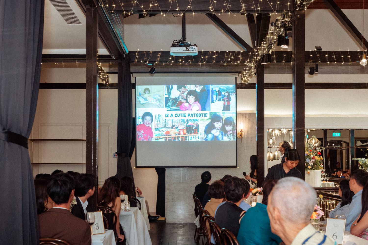 Guests seated at tables watch a photo slideshow projected on a large screen during an indoor evening event or celebration.