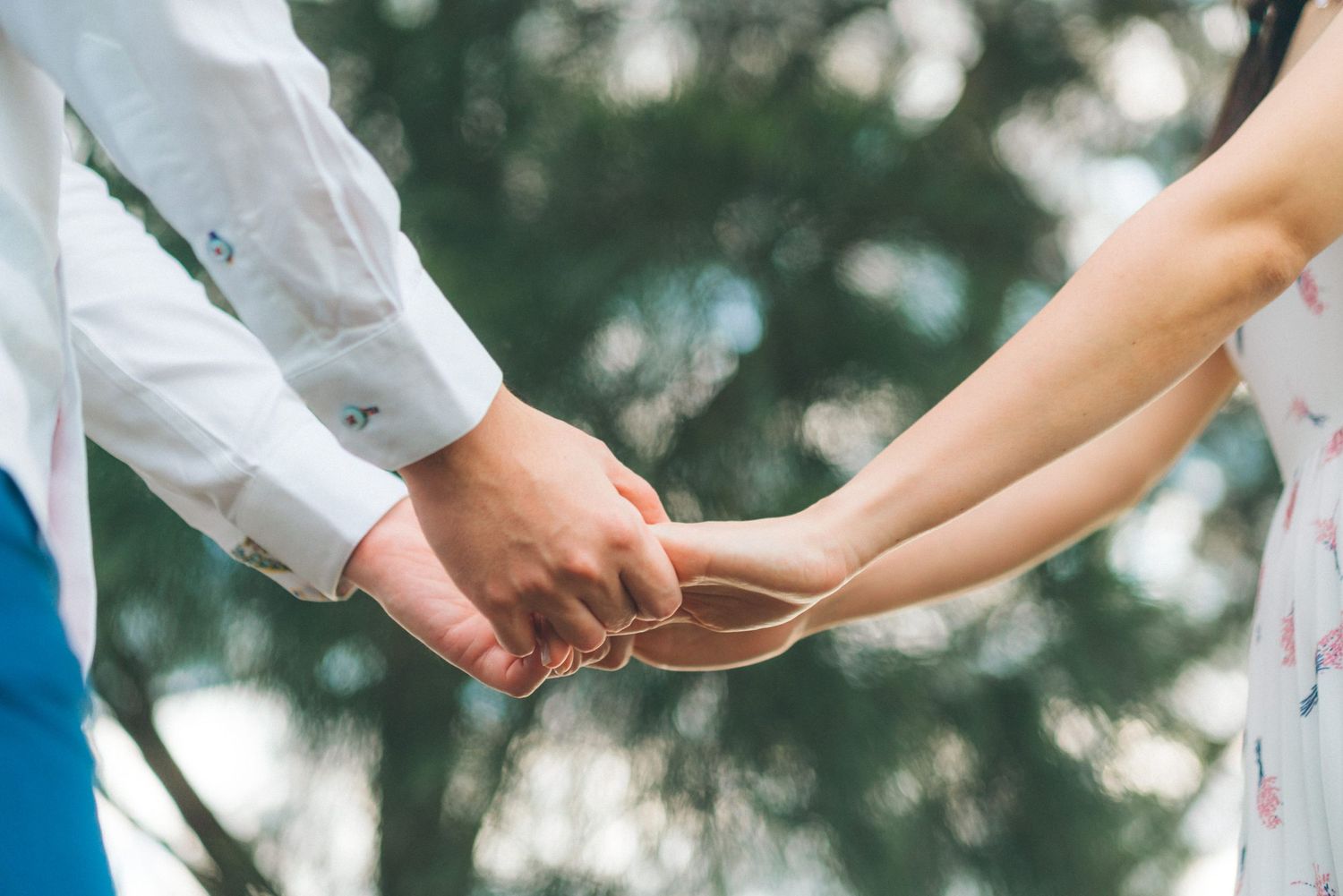 Two people in white clothing hold hands tenderly in an intimate outdoor moment with blurred green background.