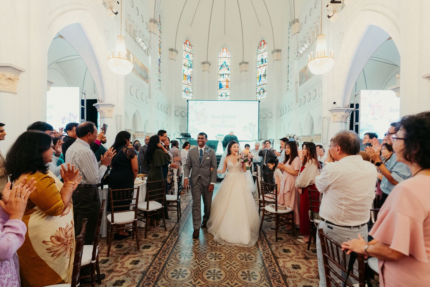 Bride and groom walk down the aisle of an elegant church as guests applaud during a beautiful wedding ceremony.