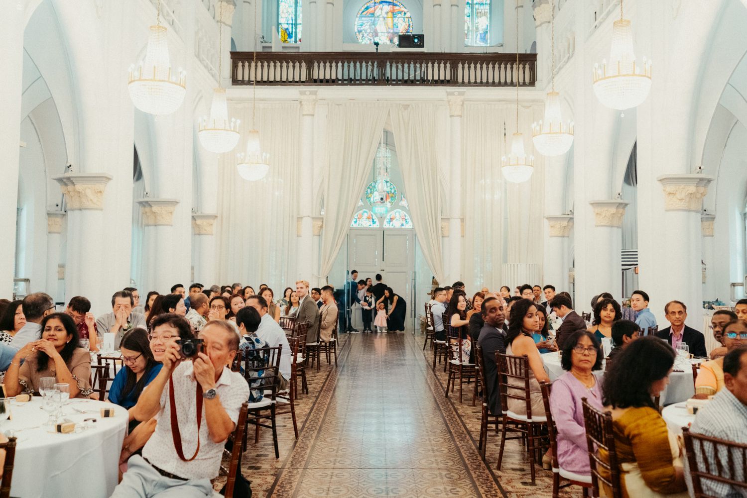 Elegant wedding reception in a grand white church hall with guests seated at decorated tables under high ceilings.