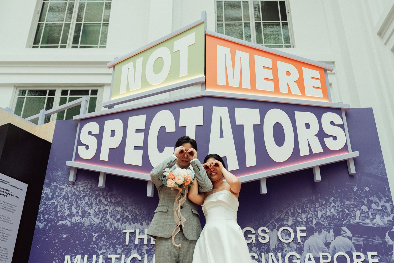 Couple posing joyfully in front of a colorful Not Mere Spectators sign at a Singapore multicultural exhibition.