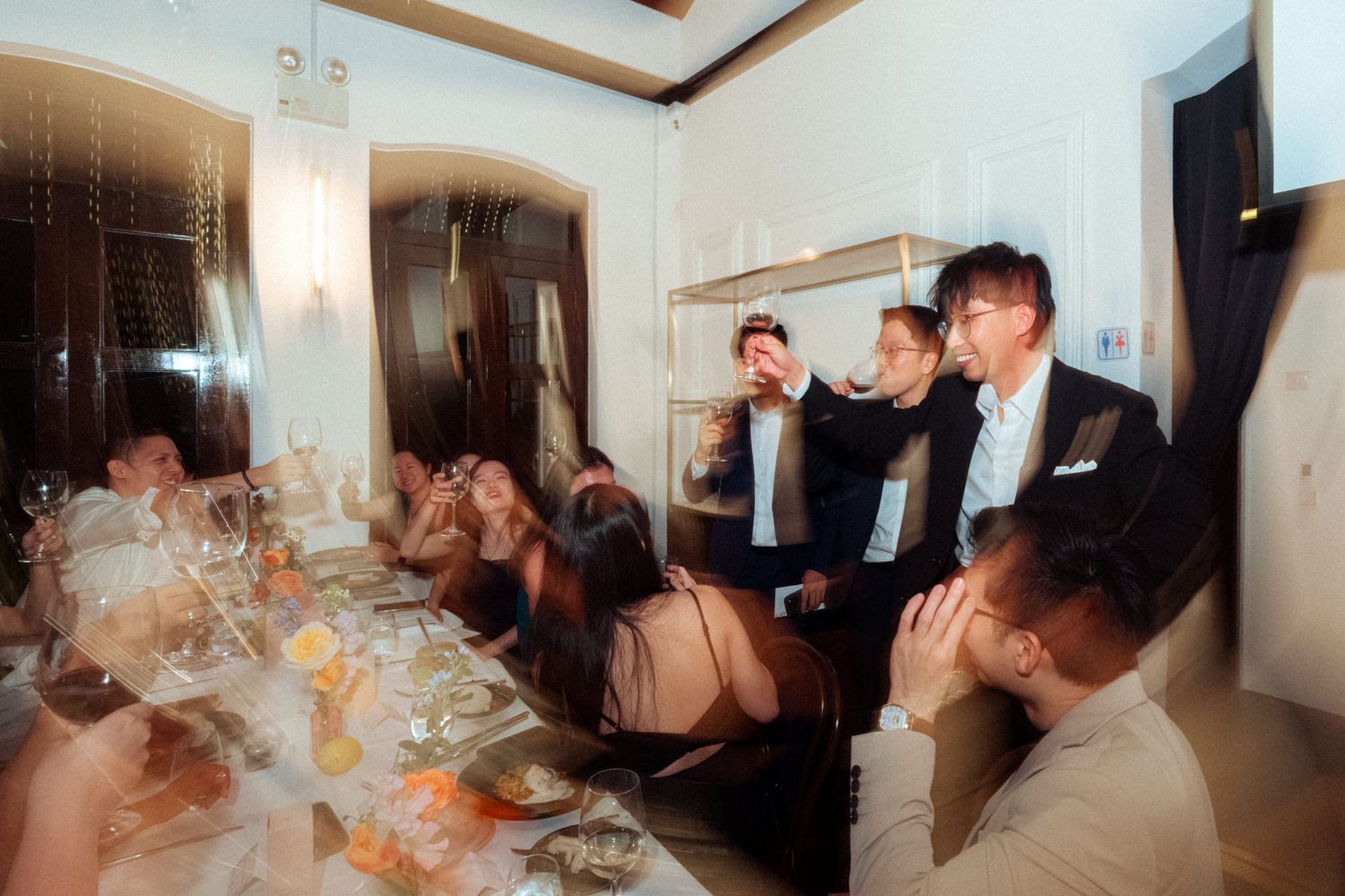 Elegant dinner party guests gathered around a long table in a luxurious dining room, toasting with drinks.