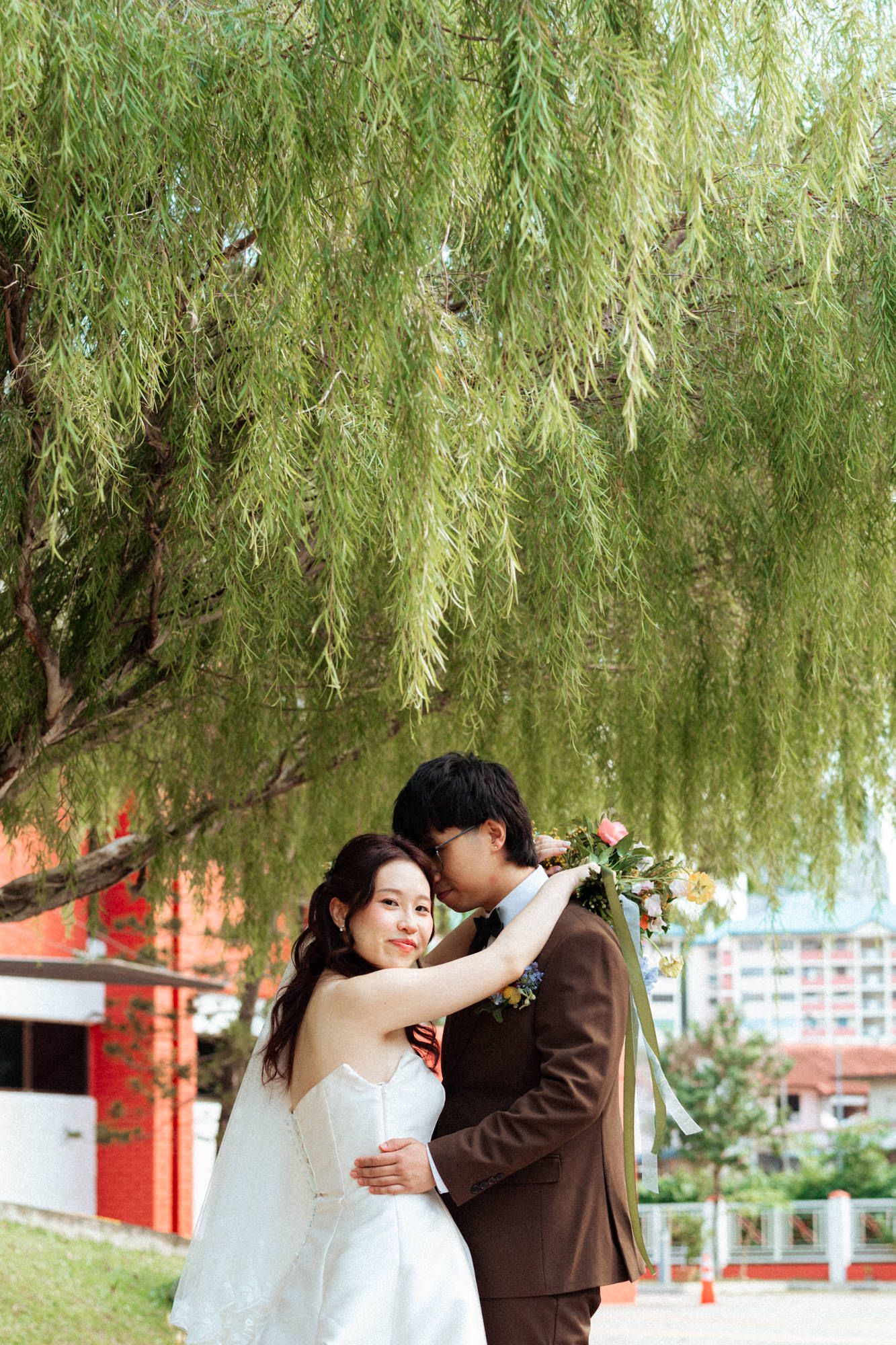 Bride and groom sharing a romantic moment under a willow tree, with the groom kissing her forehead outdoors.