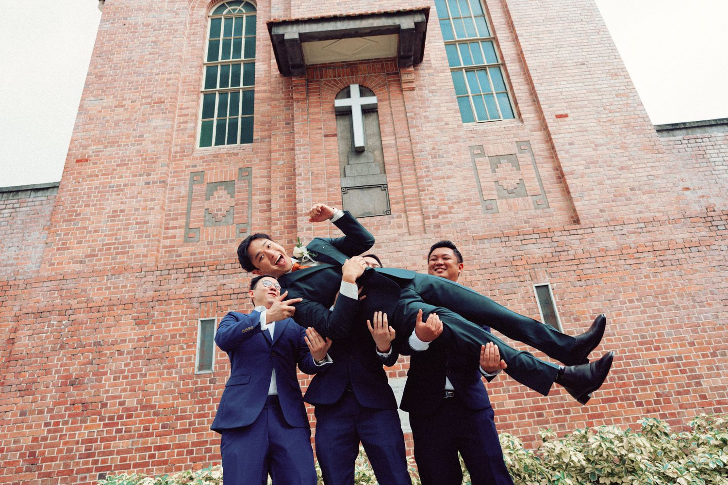 Groomsmen in navy suits lifting the groom outside Prinsep Street Presbyterian Church during a wedding celebration.