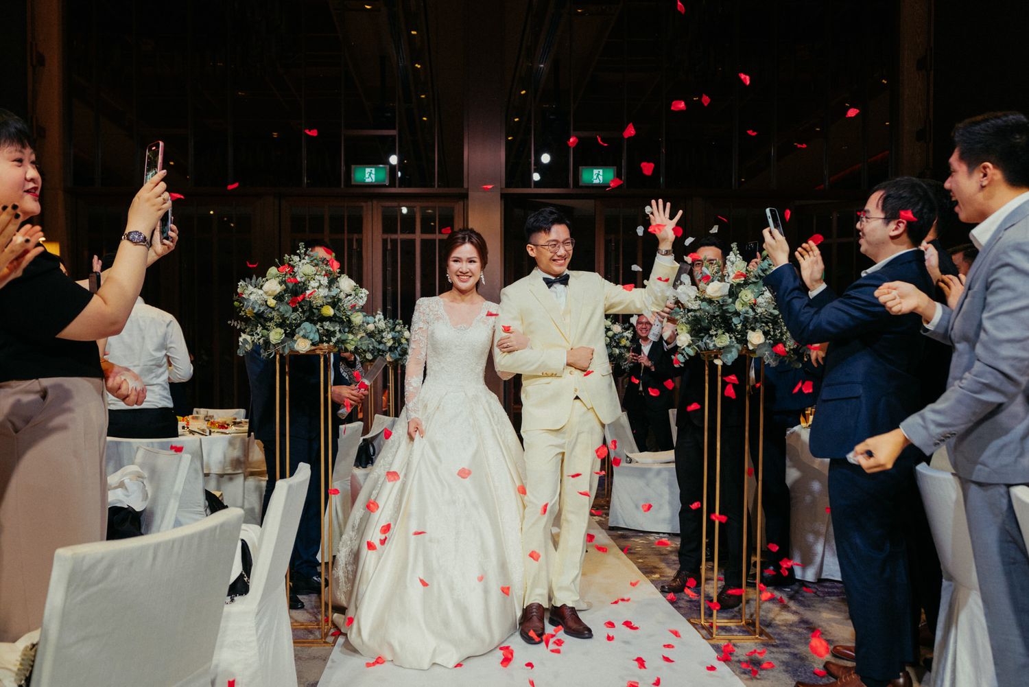 Bride and groom walk down the aisle as guests toss rose petals at their elegant indoor wedding reception.