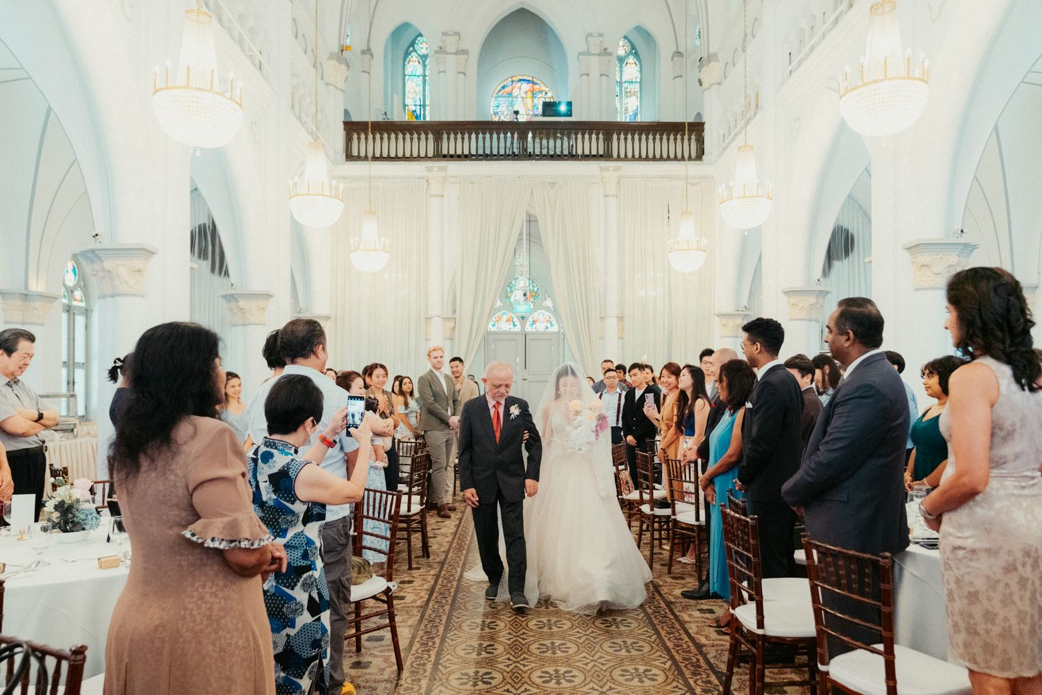 Bride walks down the aisle with her father in an elegant white church filled with wedding guests.