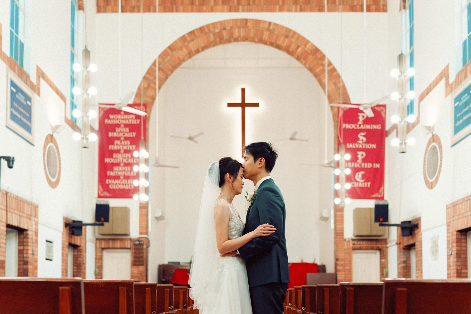 Wedding couple embraces under arched church ceiling with cross in background.