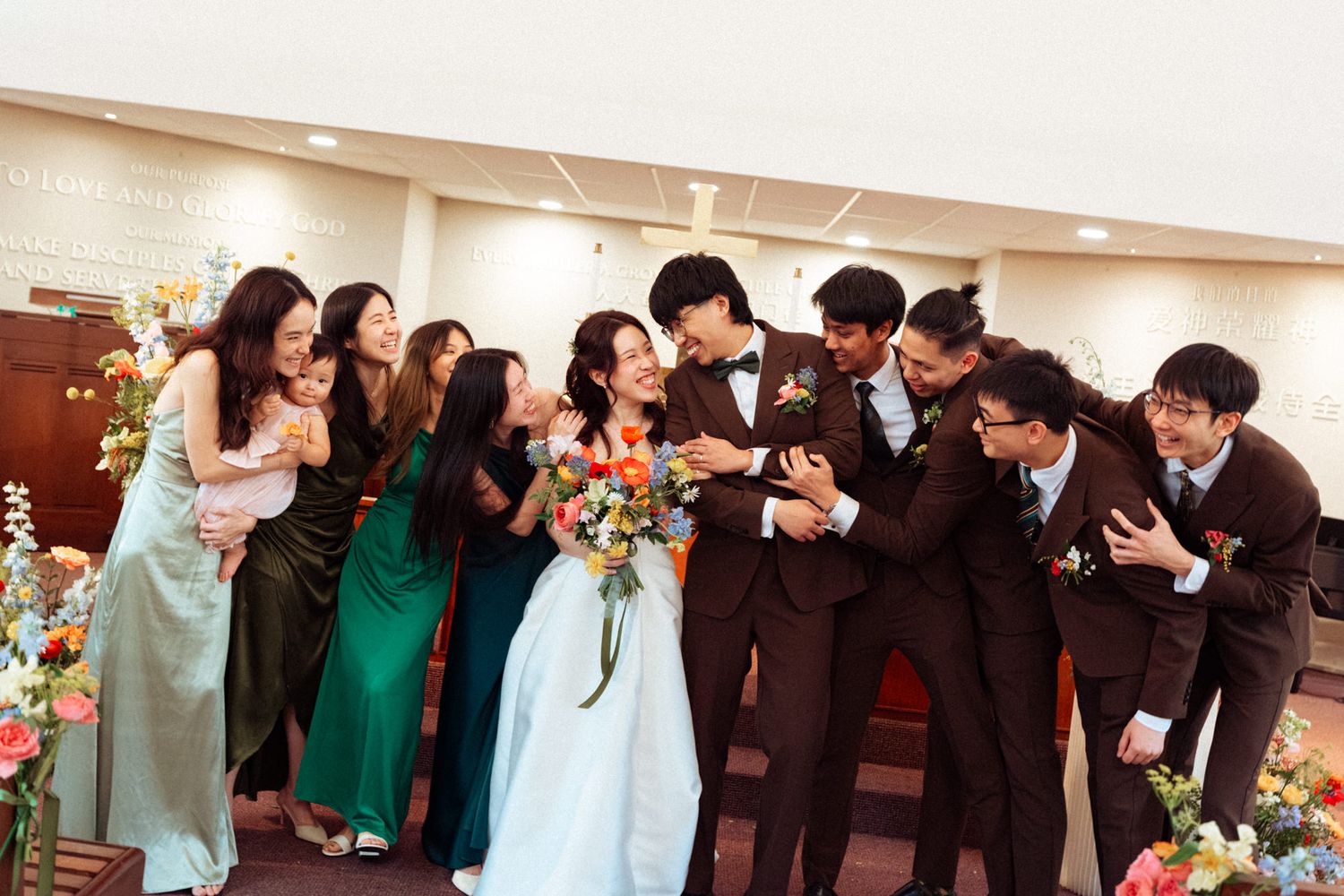Wedding party laughing together, bride in blue gown holding colorful bouquet, bridesmaids in green, groomsmen in brown suits.