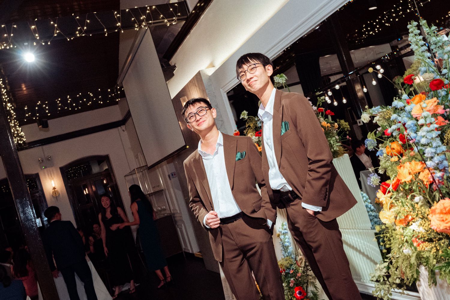 Two young men in matching brown suits smiling at an elegant event venue with colorful floral arrangements.