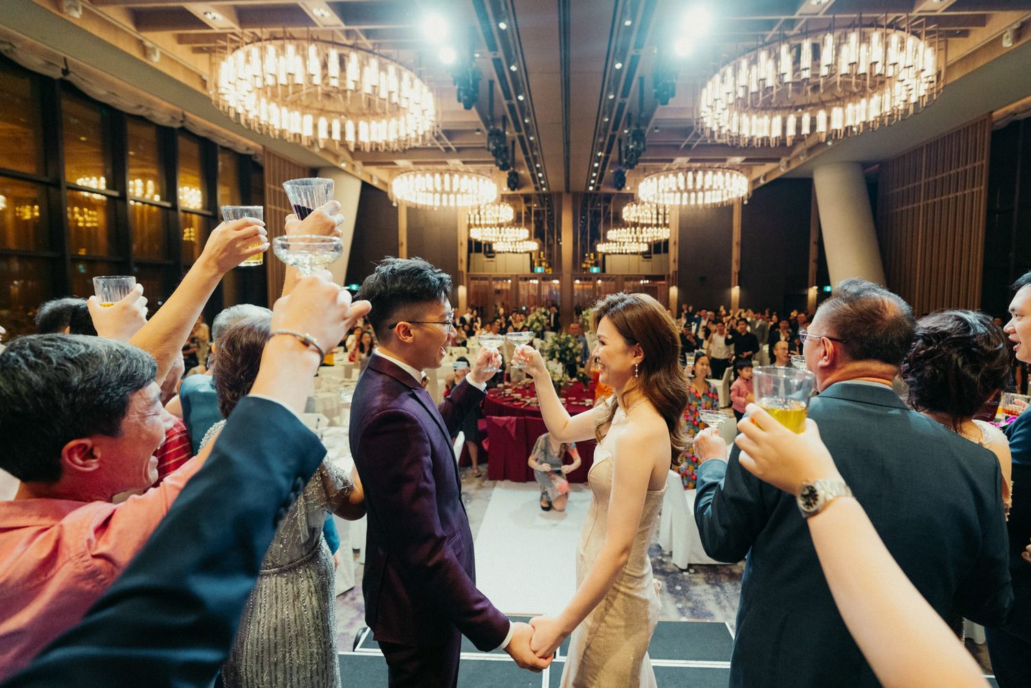 Couple holds hands during wedding reception toast in elegant ballroom with chandeliers and guests raising glasses.