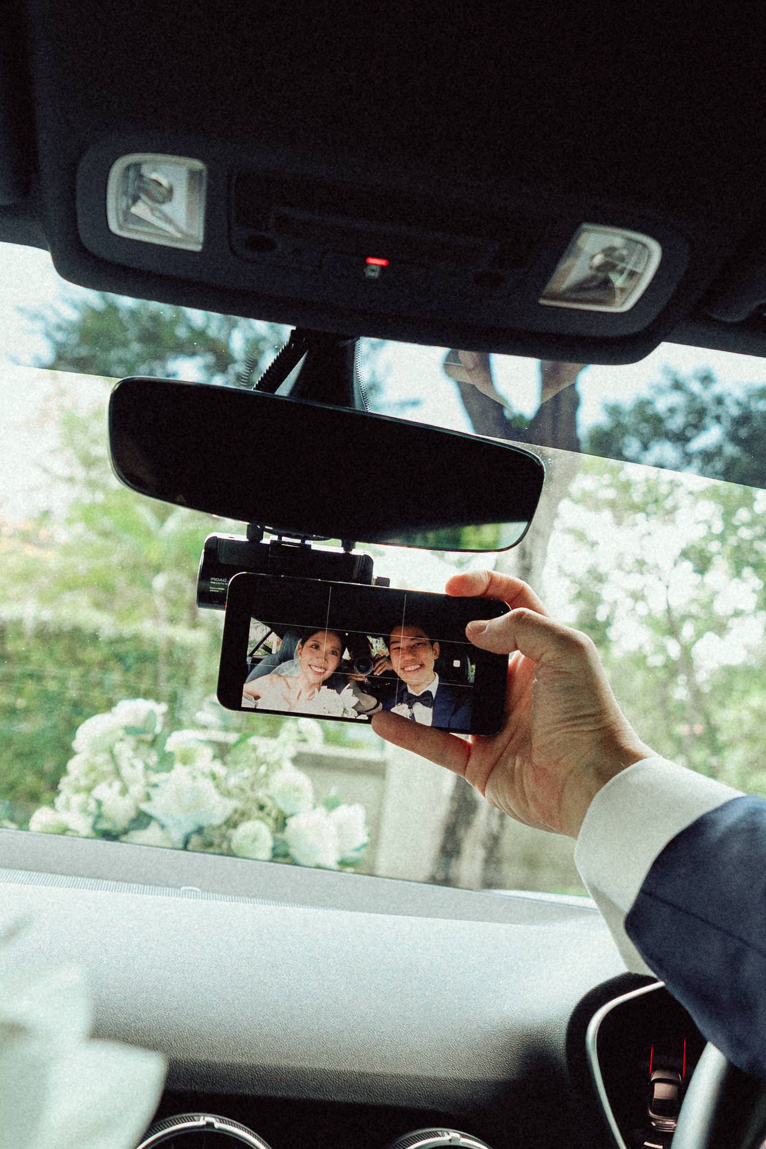 Hand holding phone to capture wedding photo through car mirror with cherry blossoms visible outside.