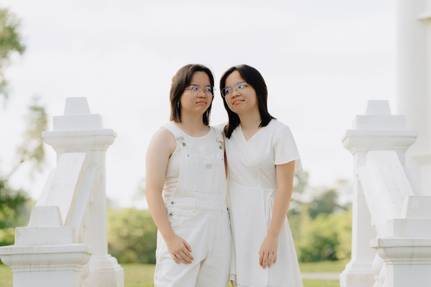 Two women in white dresses stand close together outdoors near white pillars in a romantic garden setting.
