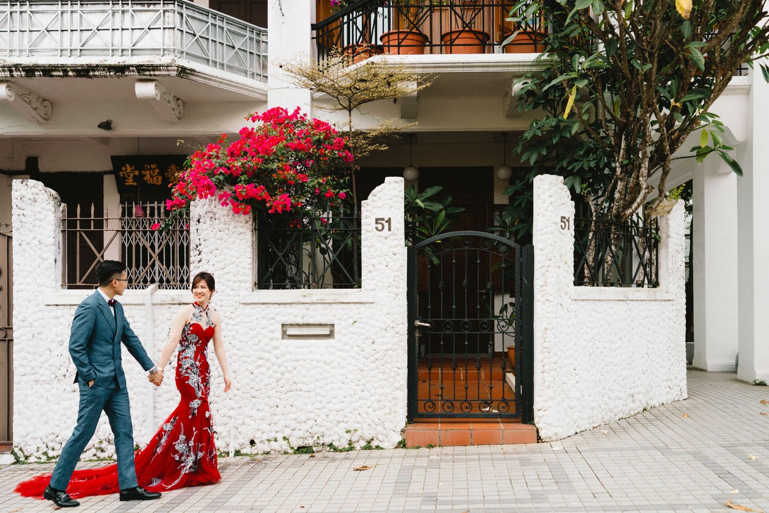 A couple shares a romantic moment in front of a white colonial building with ornate gates.