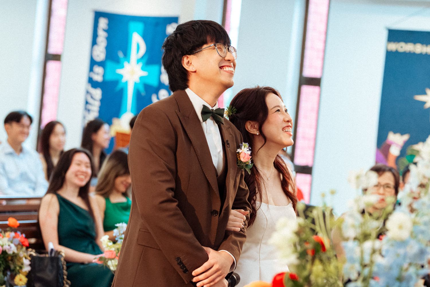 A couple in wedding attire smiles joyfully at a church ceremony, groom in brown suit, bride holding flowers.