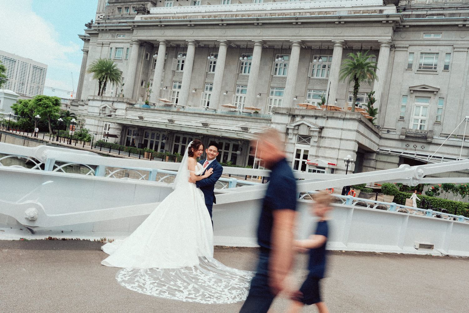 A couple in formal wedding attire poses together on a bridge with a grand architectural building in the background.