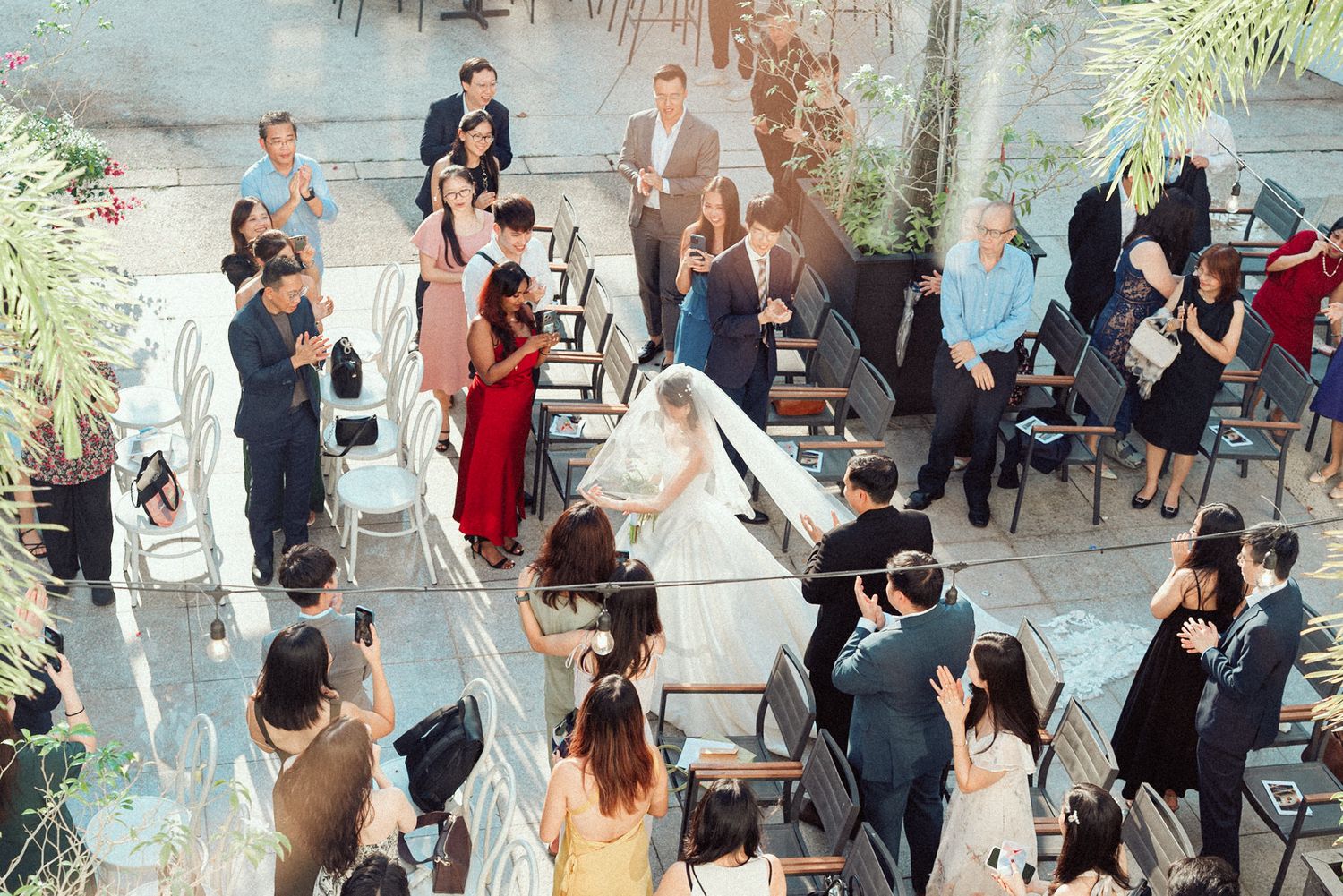 People gather at an outdoor event space with tables and chairs under string lights and palm trees.