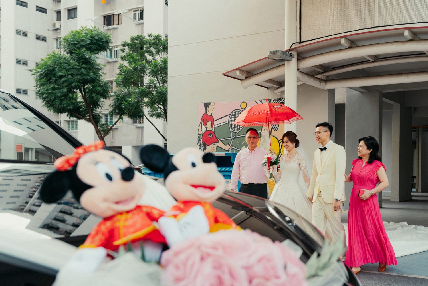 Wedding couple poses near a car decorated with Mickey Mouse plush toys, while guests hold a red umbrella outside.