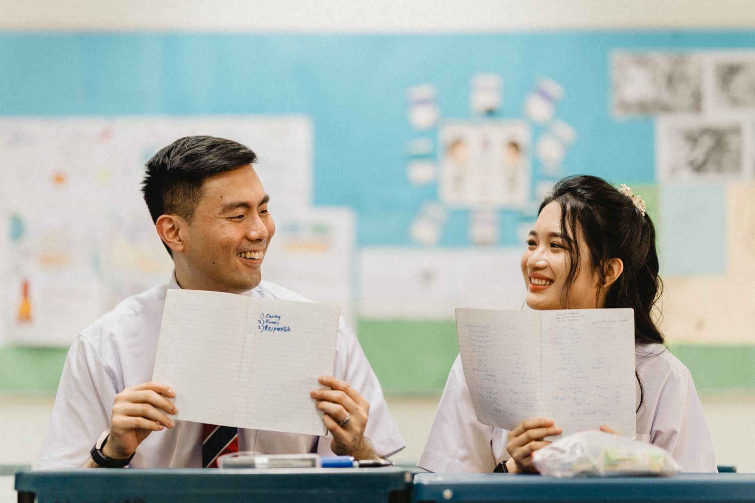 Two students in school uniforms sharing a lighthearted moment while reading papers in a classroom.