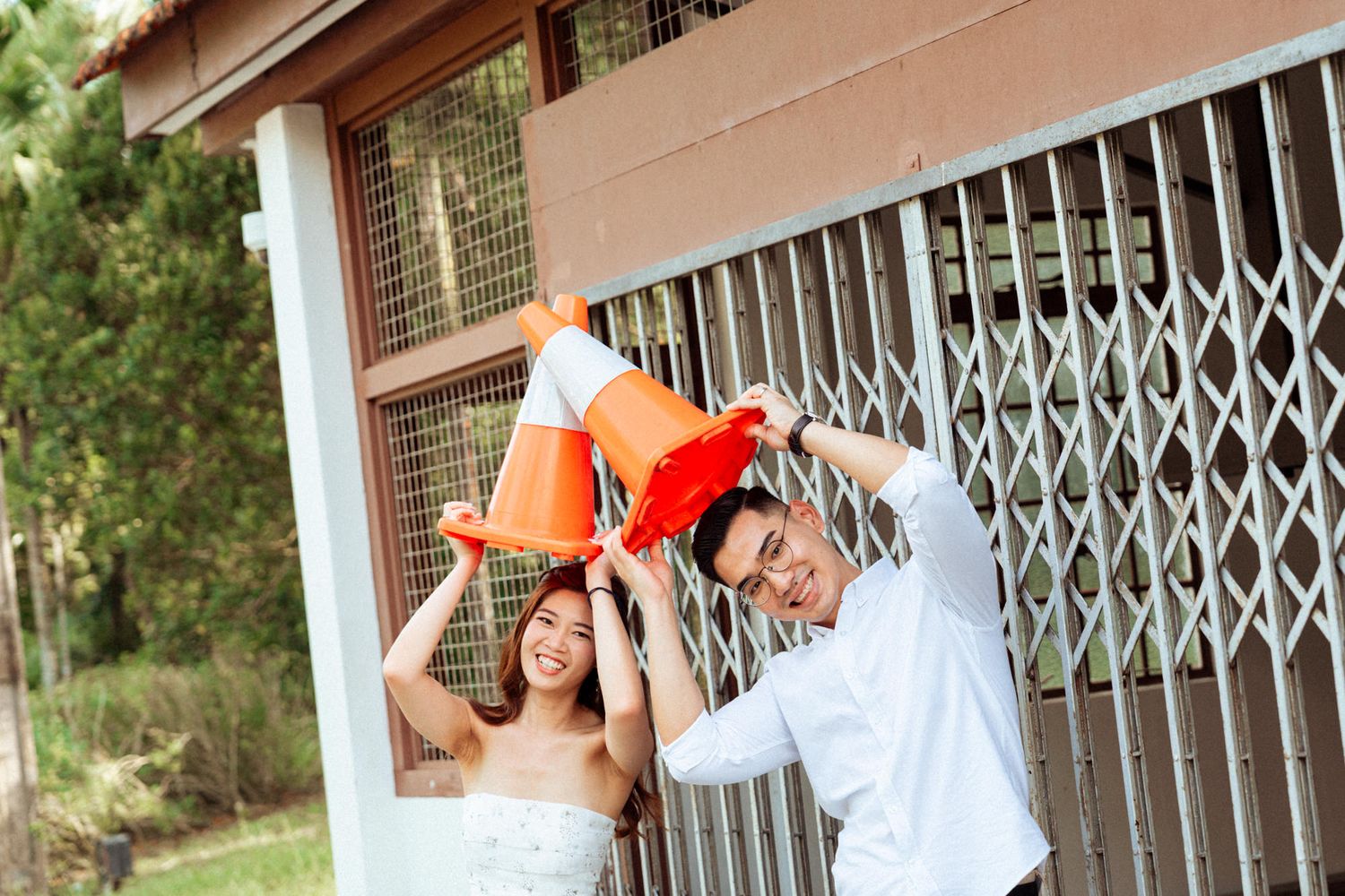 A couple in formal attire pose playfully against a white building with retractable metal gates.