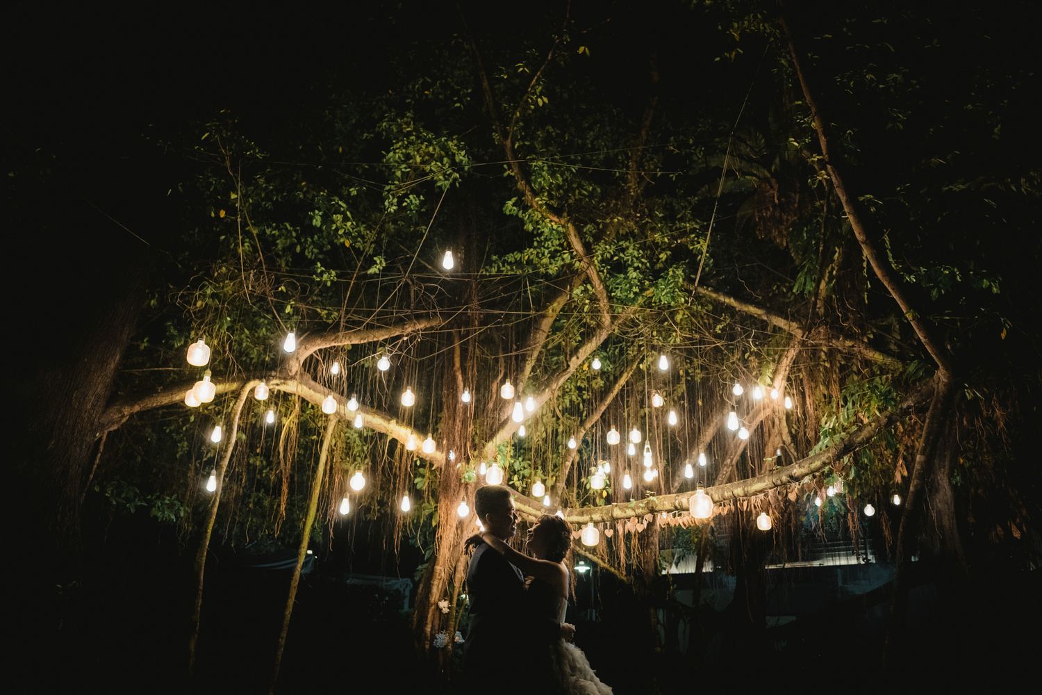 Couple sharing first dance under glowing string lights in trees at romantic outdoor night wedding reception.