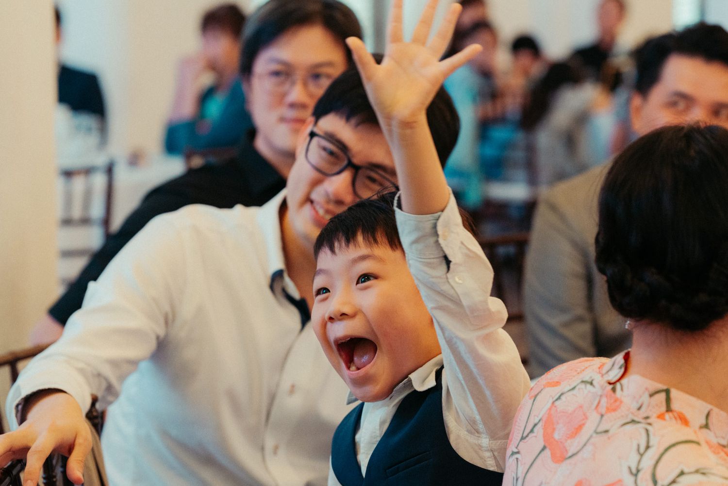 Excited young boy raises hand and laughs at a wedding reception while seated with family.