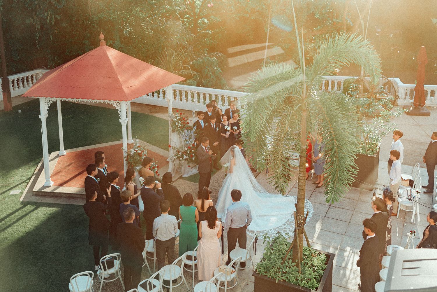 An outdoor garden wedding ceremony with a pink gazebo and guests gathered on a sunny afternoon.