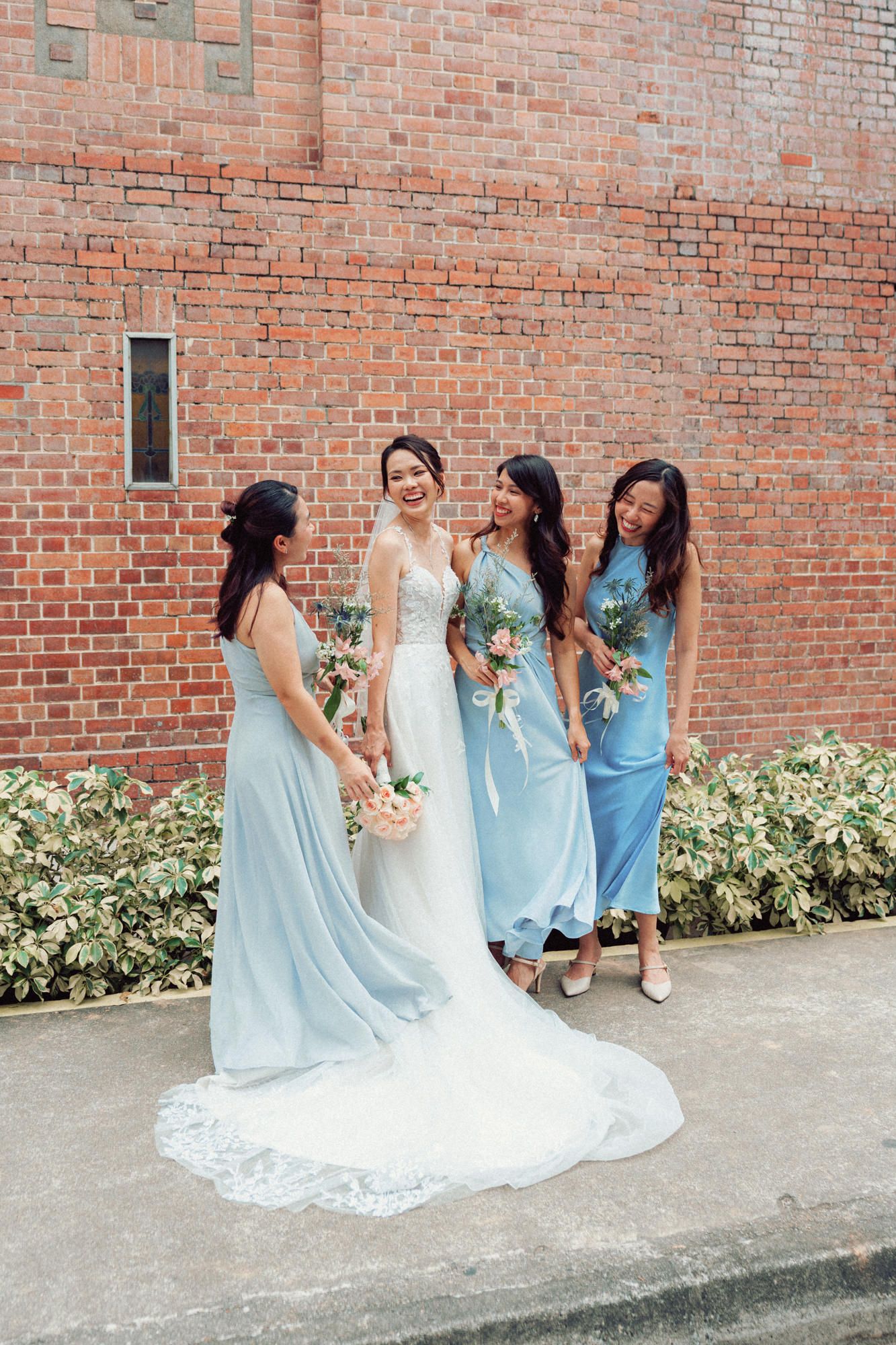 Wedding party in light blue bridesmaid dresses standing against brick wall with bouquets.