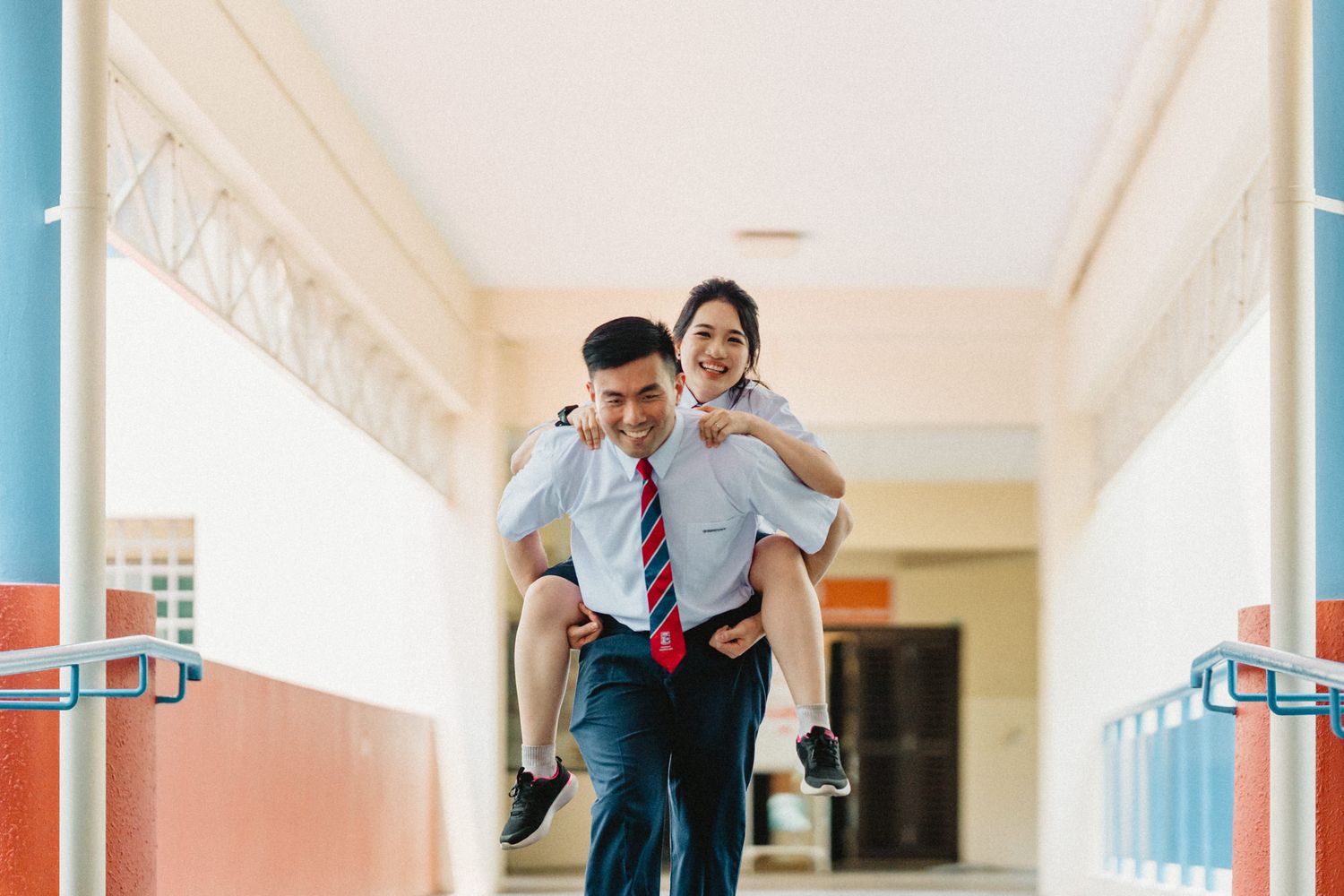 Students in school uniforms having fun in a bright hallway with one giving a piggyback ride to another.