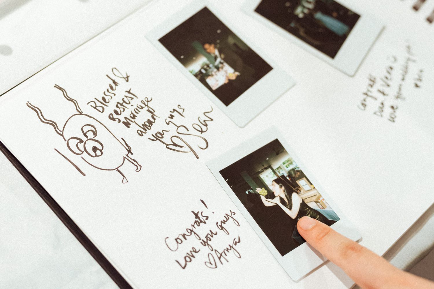 A person points to Polaroid photos and handwritten notes in a wedding guest book on a white table.