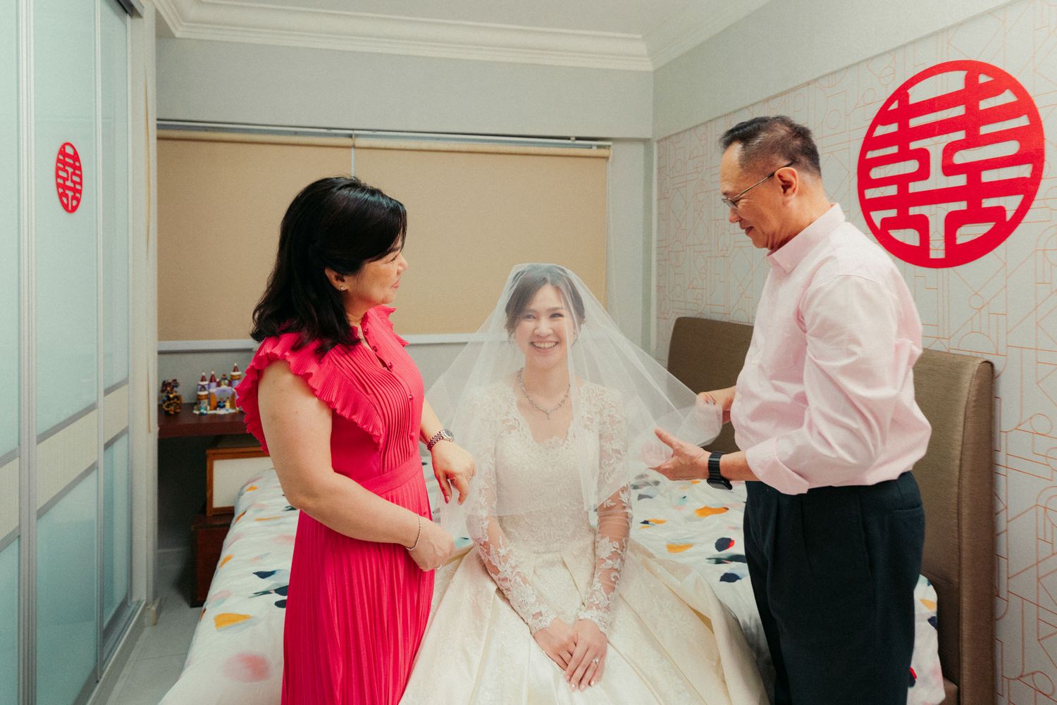 Bride in white wedding gown and veil with parents in a Chinese-decorated room with red double happiness symbol.