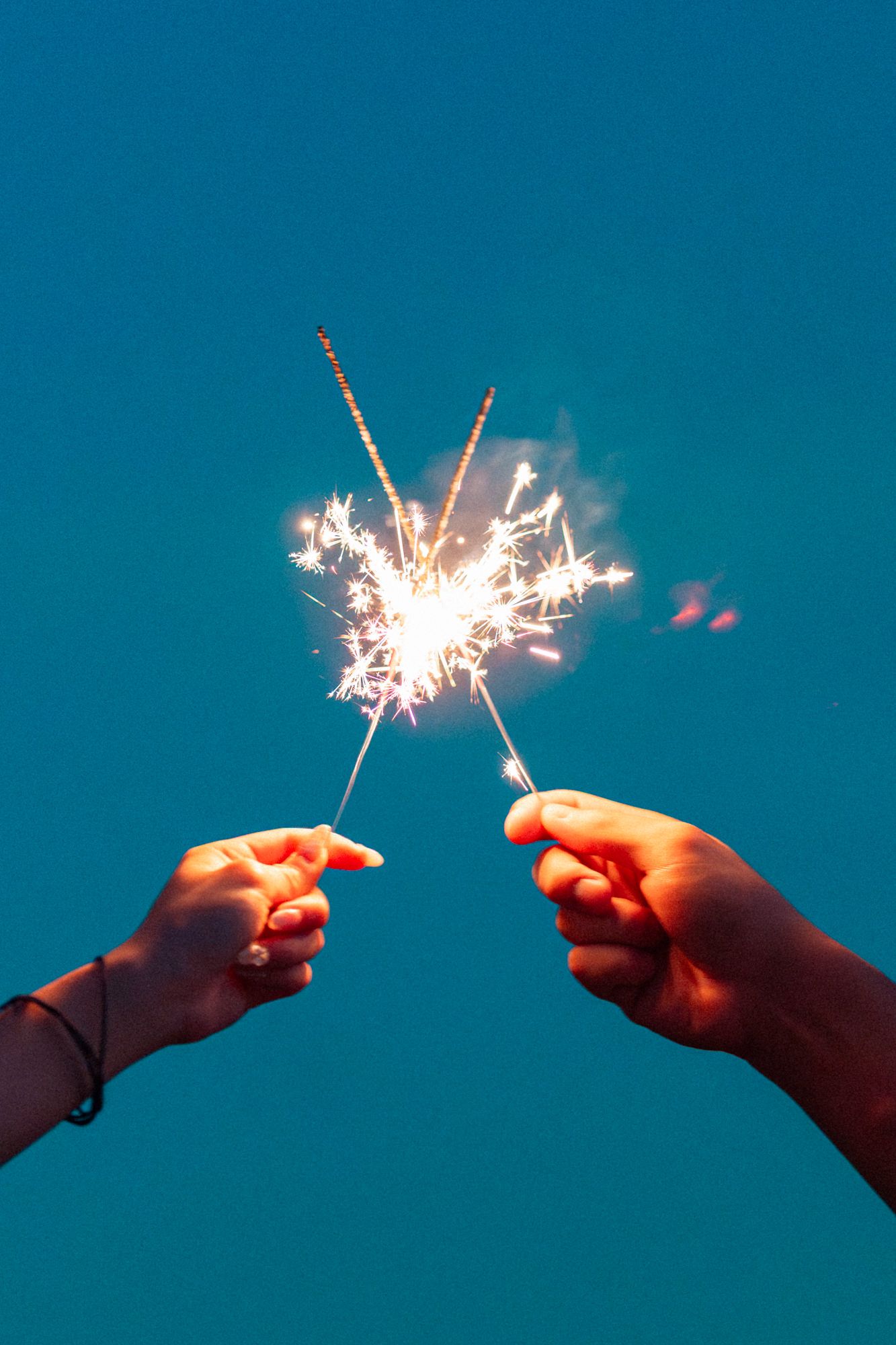 Multiple hands hold lit sparklers against a deep blue sky creating a festive celebration scene.