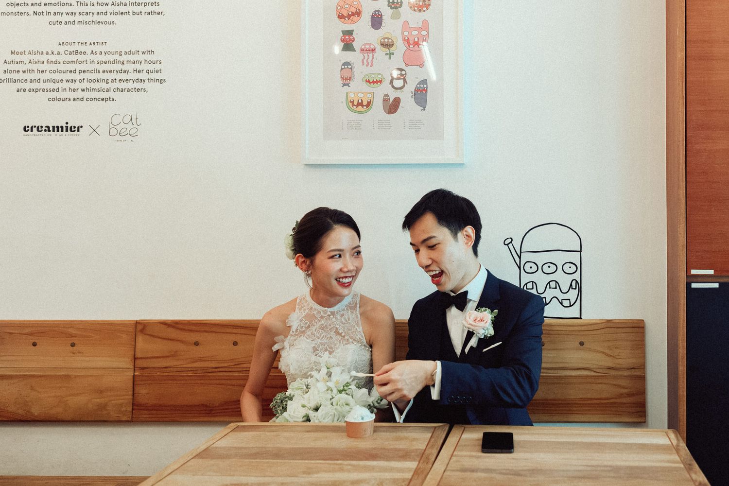 A bride and groom share a joyful moment at a wooden cafe table, with the bride in a white lace dress and groom in a navy suit.