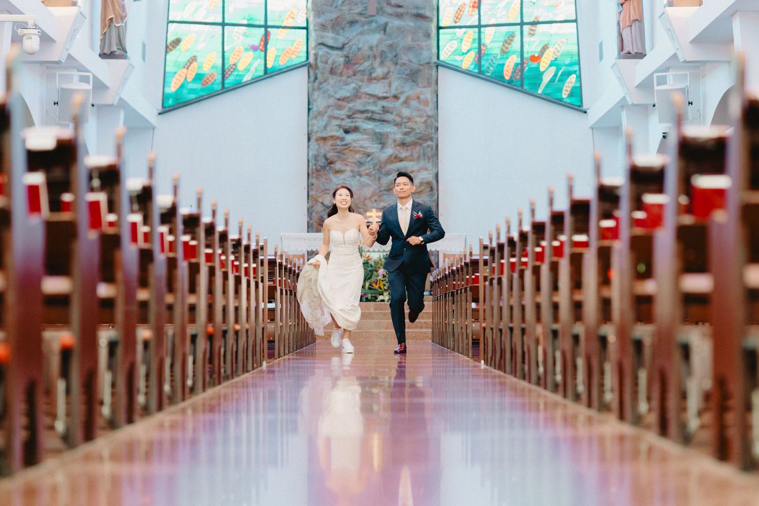 Joyful bride and groom running down church aisle after wedding ceremony, colorful stained glass windows in background.