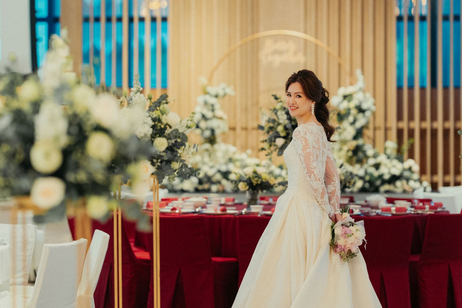 Elegant bride in a white lace gown holding a bouquet in a beautifully decorated wedding reception hall.