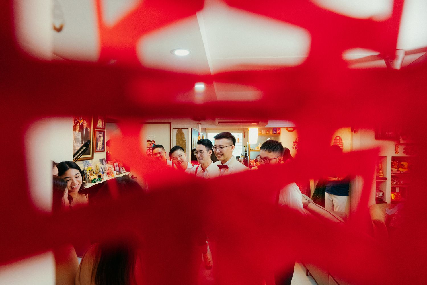 A Chinese wedding ceremony viewed through a red decorative lattice, with guests in traditional attire gathered inside.
