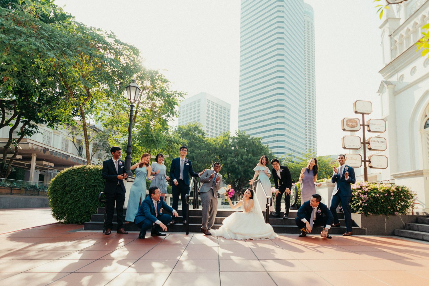Wedding party posing on sunny outdoor steps near modern skyscrapers, bride in white gown surrounded by guests.