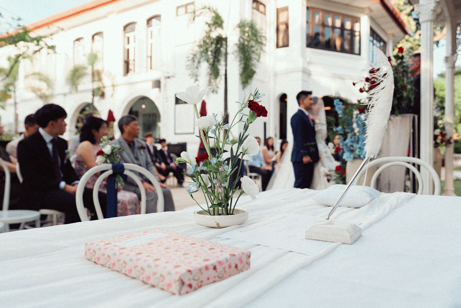 Wedding guests gather in an elegant white reception hall decorated with palm trees and floral arrangements.