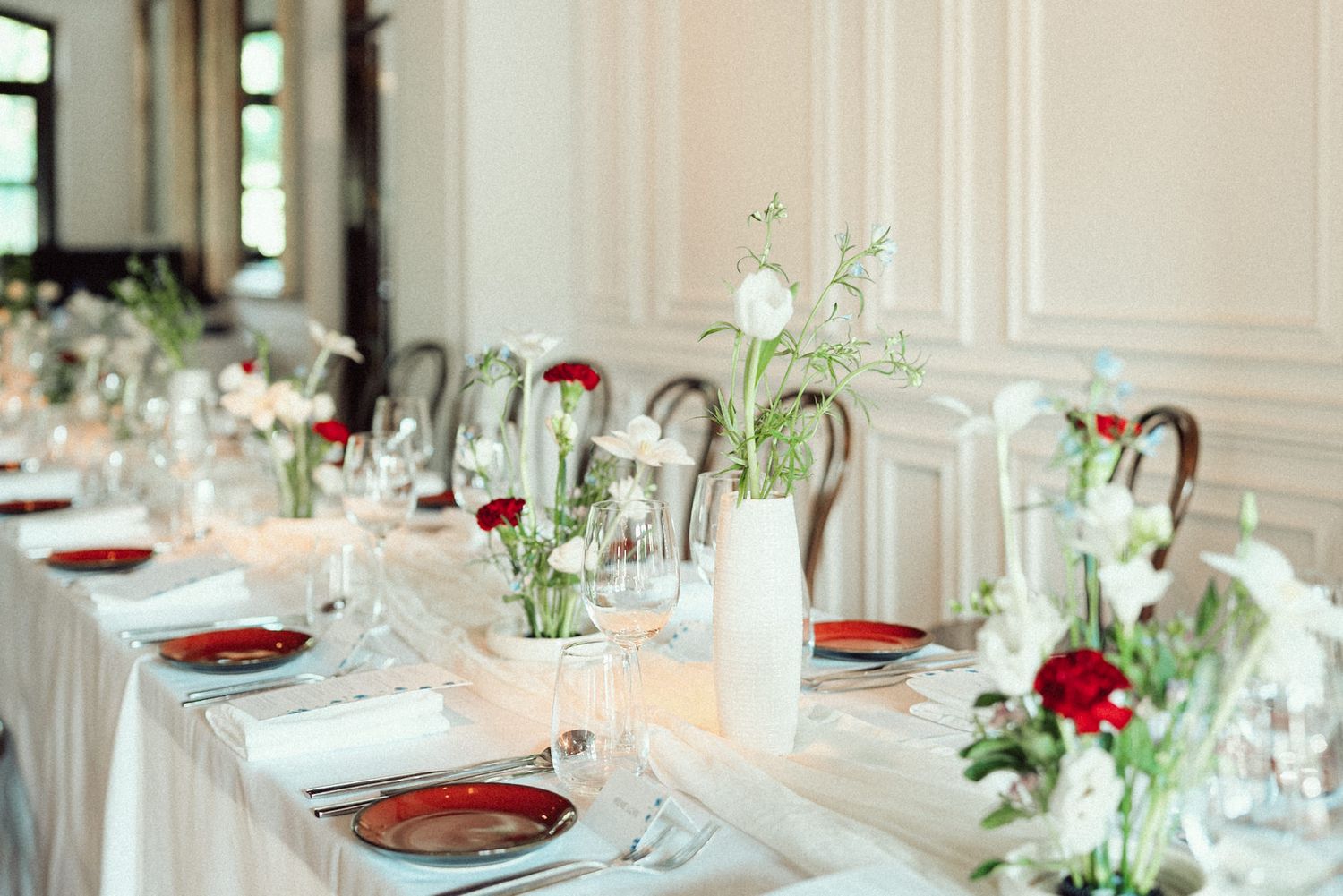 Elegant wedding reception tables with white linens, red accents, and floral centerpieces in a bright banquet hall.