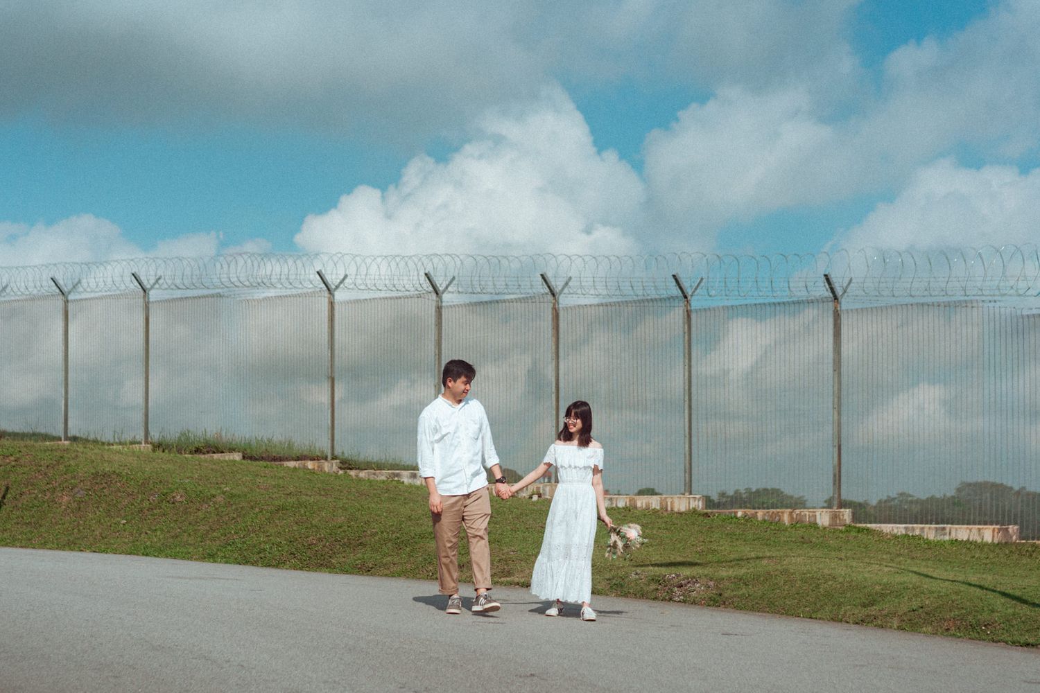 A couple walks along a grassy path beside a security fence under a cloudy sky.