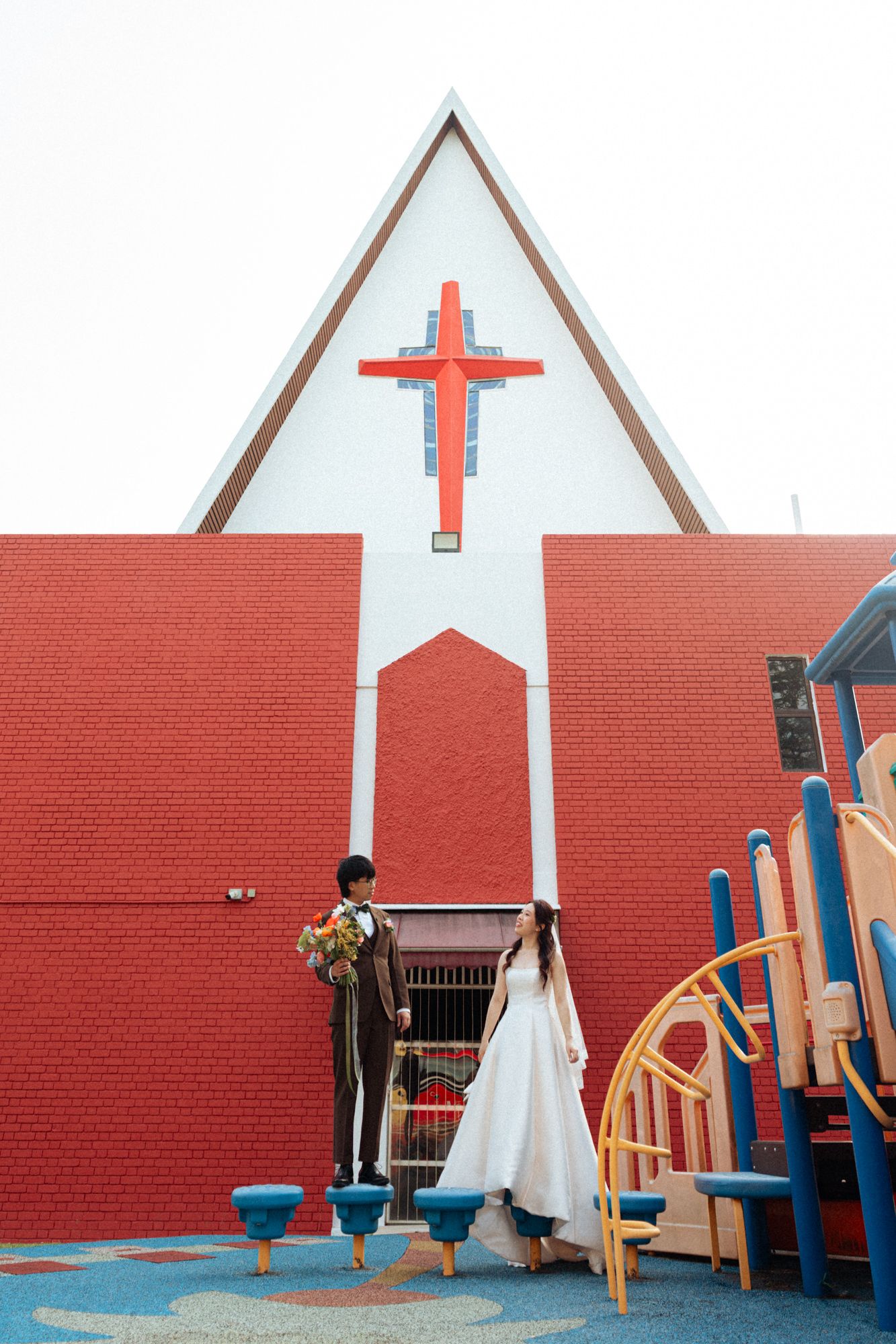 Bride and groom standing before a red brick church with a cross, next to a colorful playground.