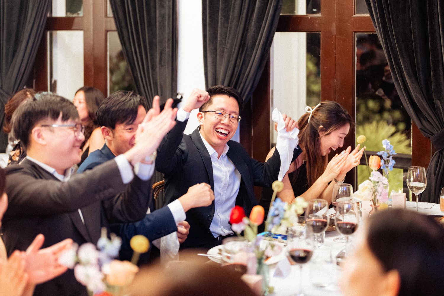 Joyful guests laughing and celebrating at an elegant dinner party with wine glasses and floral centerpieces.