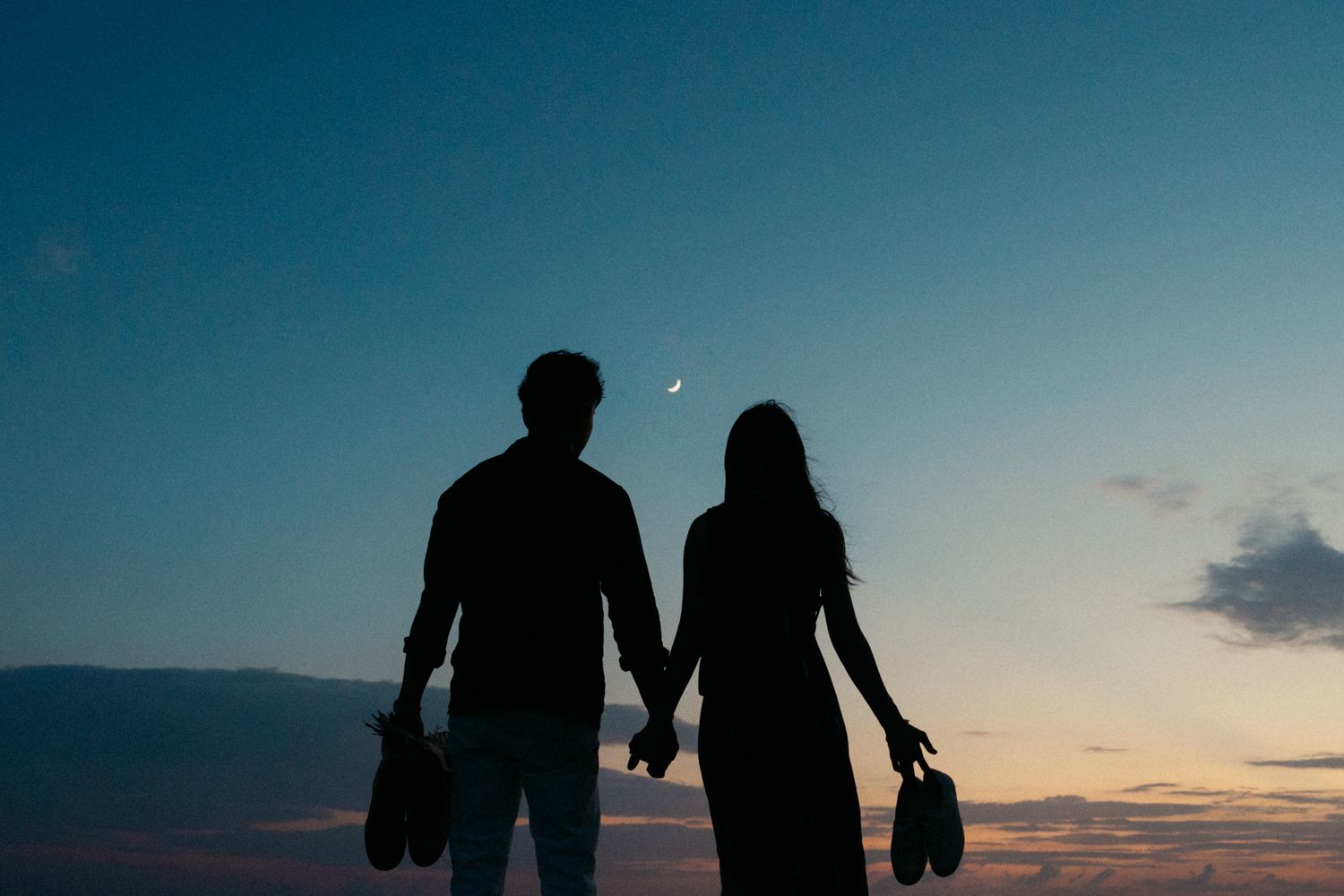 Couple's silhouettes against dramatic sunset sky at beach with cityscape in distance.