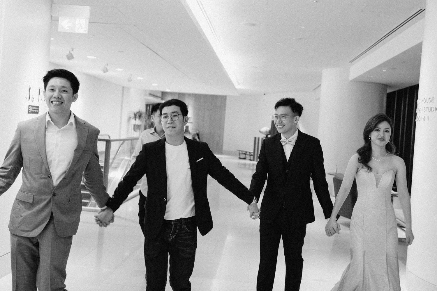 Four elegantly dressed people holding hands and walking together in a modern venue in a black and white photo.