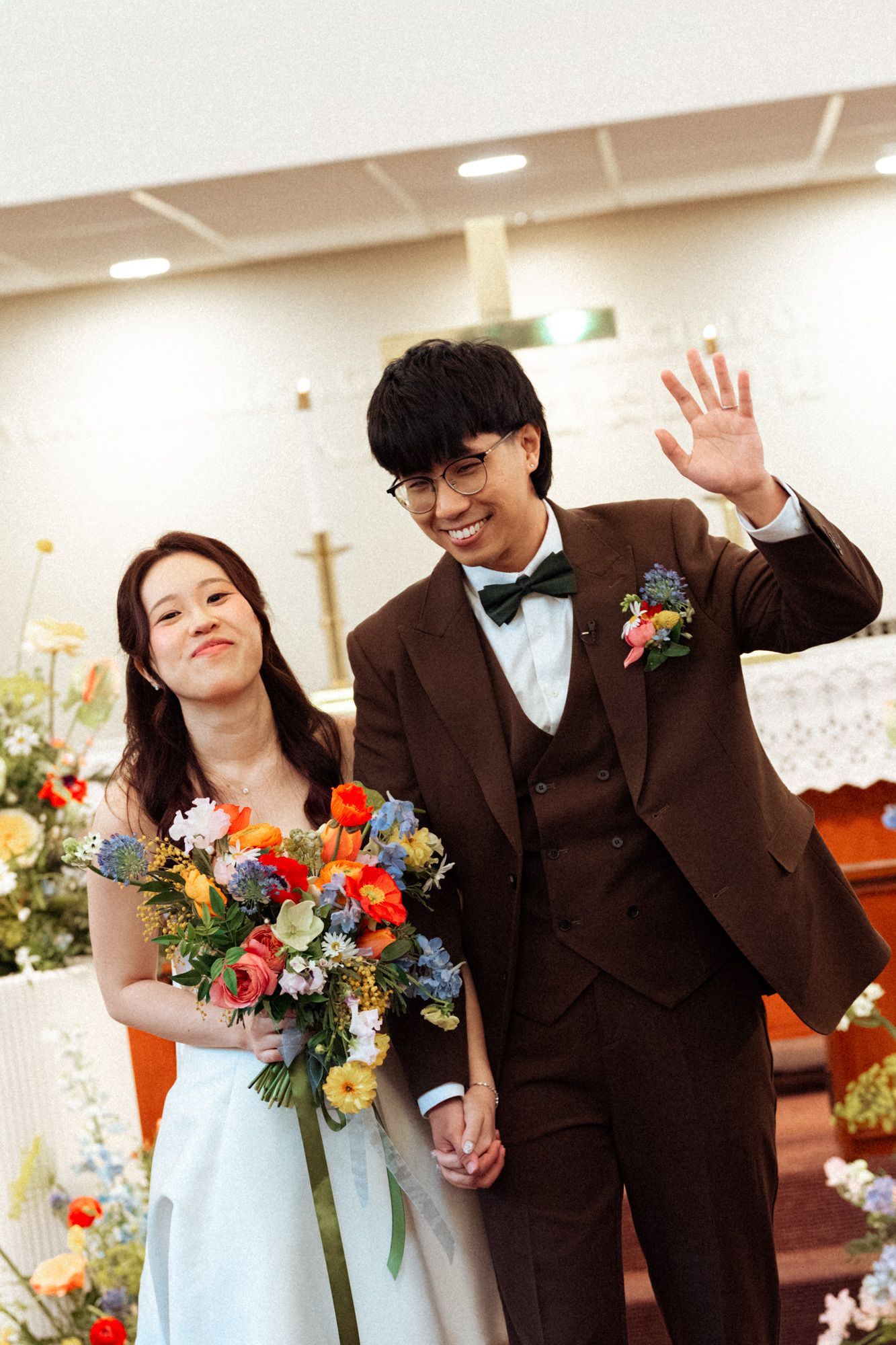 A smiling bride holding a colorful bouquet and groom in brown suit waving at their wedding ceremony in a church.