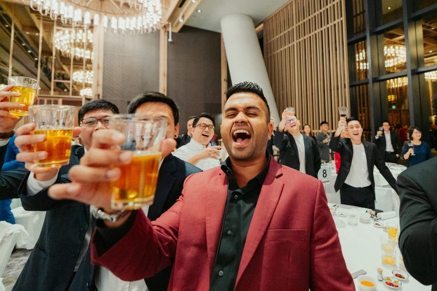 Man in red blazer laughing and toasting with beer at a lively indoor celebration event.