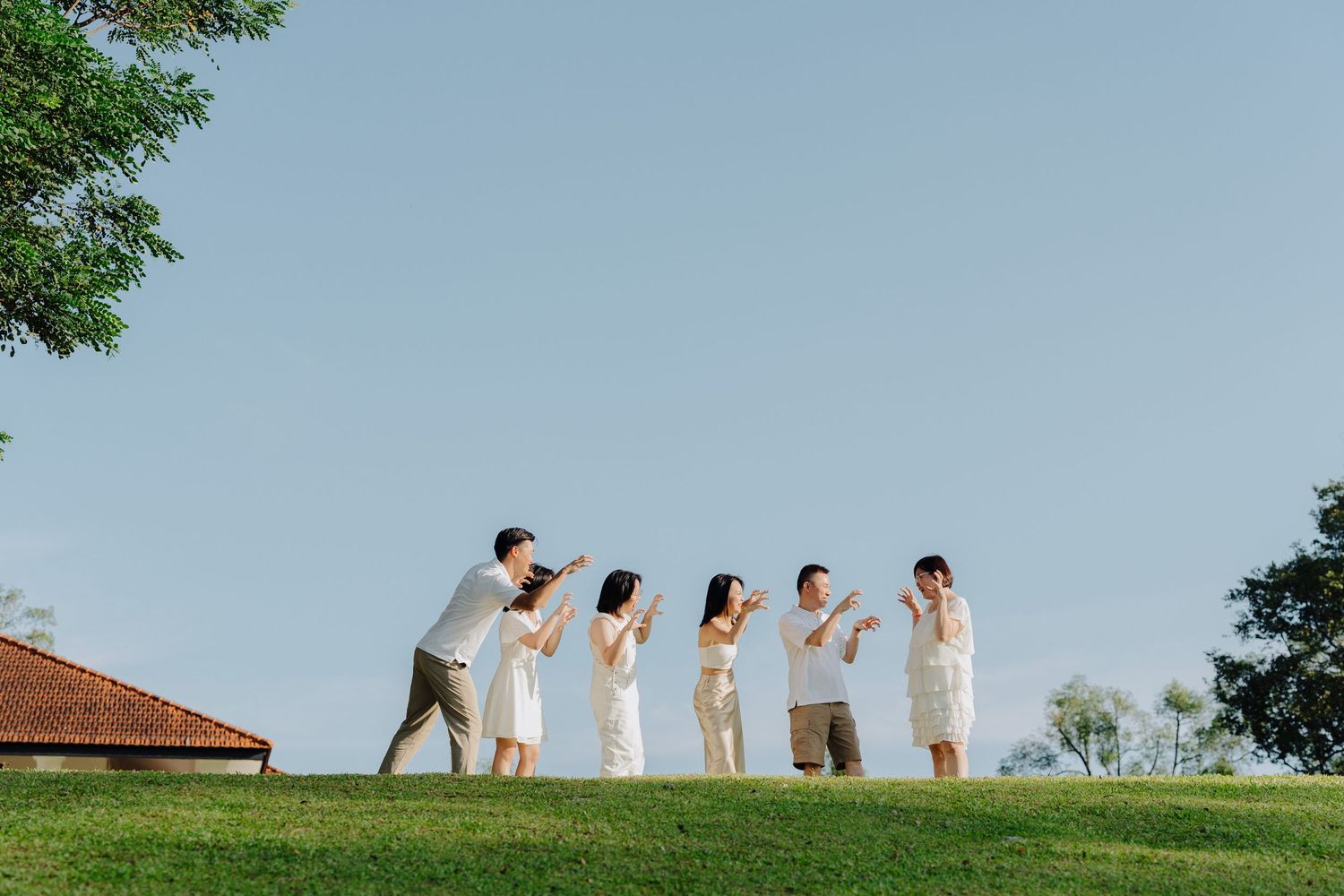 A wedding party in white attire poses together on a grassy hilltop against a bright blue sky.