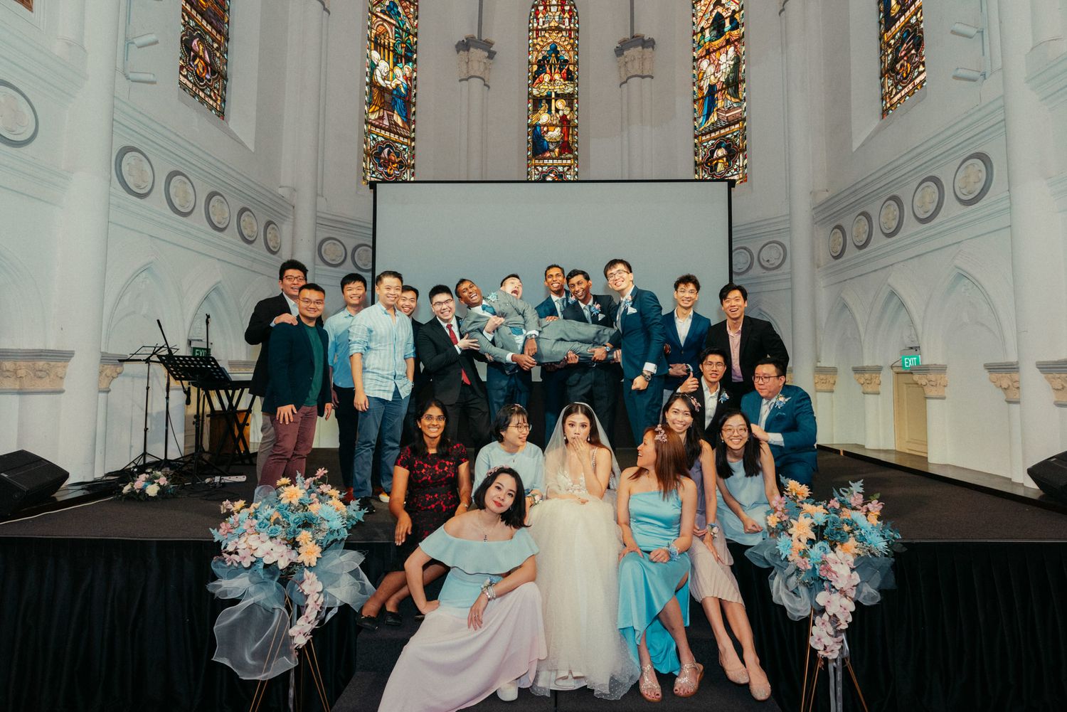 Wedding party poses in elegant church venue with stained glass windows, bride in white gown surrounded by guests in blue attire.
